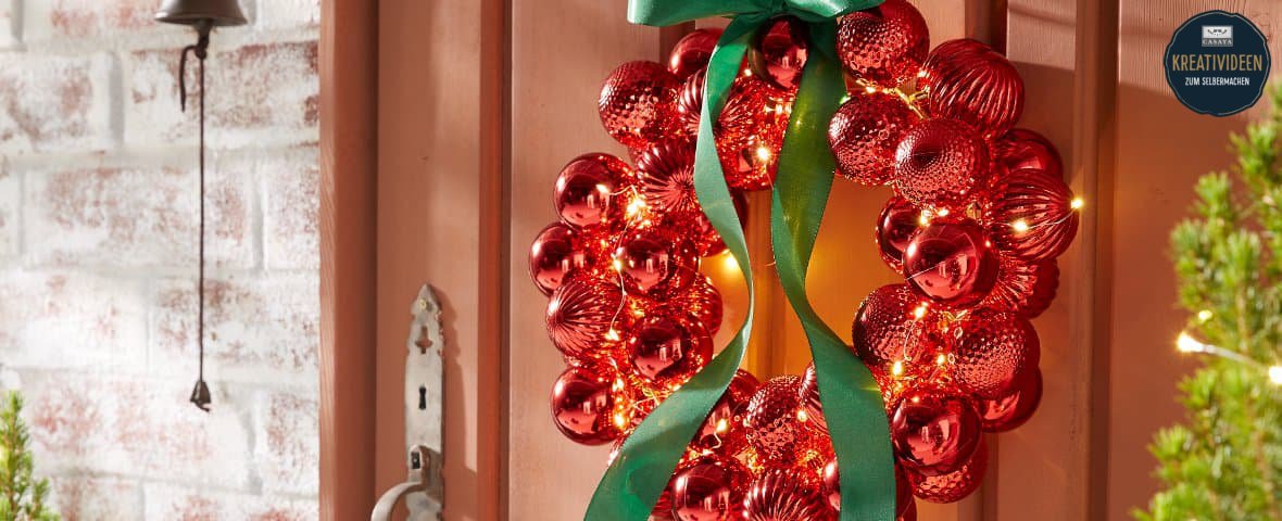 Ein Türkranz aus roten glänzenden und strukturierten Weihnachtskugeln hängt an einer Holzhaustür. In den Kranz ist eine warmweiße Lichterkette eingearbeitet, oben ist er mit einer großen grünen Schleife befestigt. Oben rechts ist ein schwarzer Button auf dem das Casaya Logo abgebildet ist. Darunter steht in gelber und weißer Schrift KREATIVIDEEN ZUM SELBERMACHEN