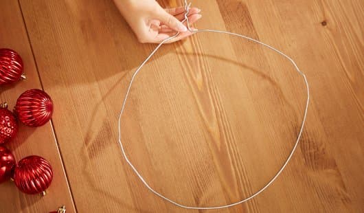 Eine Hand formt auf einem Holztisch ein Stück silbernen Draht zu einem Ring und verdrillt die Enden. Links im Bild liegen rote strukturierte Weihnachtskugeln bereit