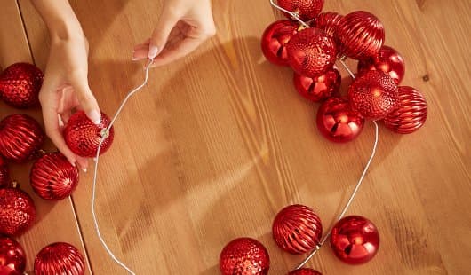 Zwei Hände fädeln abwechselnd glänzende und strukturierte rote Weihnachtskugeln auf einen silbernen Draht, der zu einem offenen Ring geformt ist. Die Kugeln liegen in Gruppen auf einem Holztisch bereit