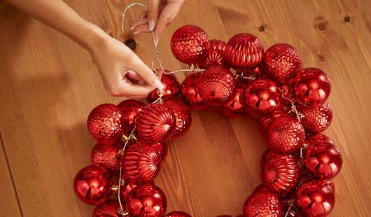 Zwei Hände verschließen den mit roten Weihnachtskugeln bestückten Drahtkranz, indem sie die Drahtenden miteinander verdrehen. Zwischen den Kugeln ist eine kleine Lichterkette sichtbar, der Kranz liegt auf einem Holztisch