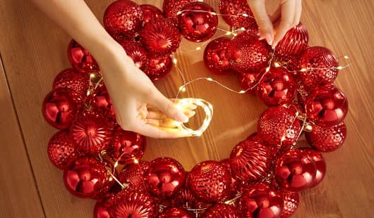 Zwei Hände legen eine kleine warmweiße Lichterkette um einen mit roten Weihnachtskugeln gestalteten Kranz. Die Lichterkette wird locker zwischen den Kugeln verteilt, der Kranz liegt auf einem Holztisch