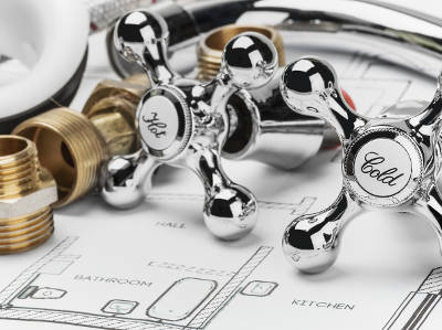 Zwei klassische Armaturengriffe für Warm- und Kaltwasser liegen auf einem Badezimmergrundriss. Umgeben sind sie von weiteren Sanitärteilen wie Schläuchen und Gewindemuffen