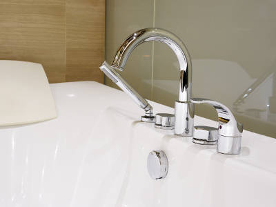 Eine moderne Badewanne mit glänzender Chromarmatur und Handbrause ist in einem stilvollen Badezimmer installiert. Im Hintergrund sind helle Wandverkleidungen aus Holz und Glas zu sehen