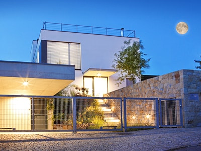 Ein modernes, kubisches Wohnhaus bei Nacht. Der Eingangsbereich ist hell beleuchtet. Vor dem Haus befindet sich ein Metallzaun mit Tor, daneben eine Natursteinmauer.