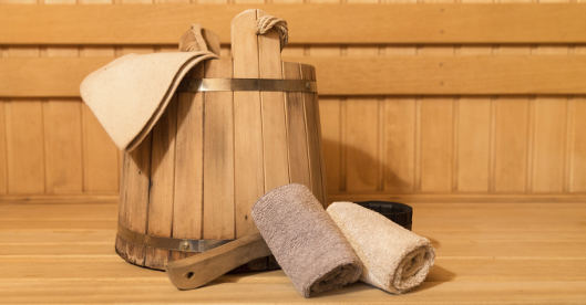 Saunazubehör mit Holzeimer, Handtüchern und Aufgusskelle auf Holzbank.