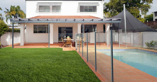 Garten mit einem rechteckigen Swimmingpool neben einer Terrasse mit Terrakottafliesen, der Pool ist durch einen transparenten Zaun von der Rasenfläche getrennt, im Hintergrund steht ein Haus