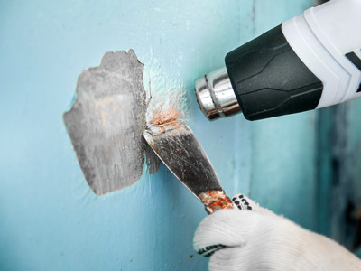 Foto einer Wand, an der mit einem Spachtel und einer Heißluftpistole blaue Farbe gelöst und abgekratzt wird