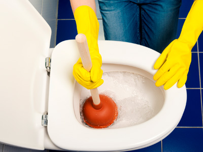 Eine Person mit gelben Gummihandschuhen verwendet einen Pümpel, um eine verstopfte Toilette zu reinigen. Die WC-Brille ist geöffnet, der Boden ist mit blauen Fliesen ausgelegt