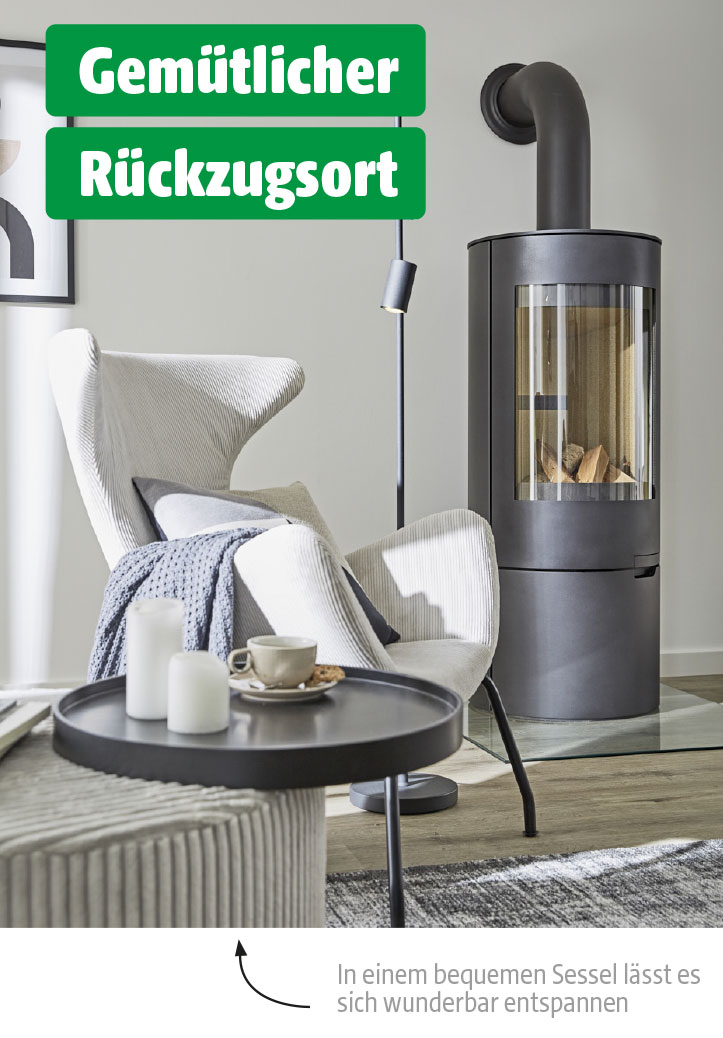 Heller Sessel mit Kissen und Decke neben einem runden Beistelltisch mit Kerzen und Tasse – vor einem Kaminofen in ruhiger Sitzecke.