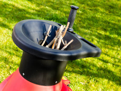 Mehrere trockene Äste stecken in der Einfüllöffnung eines roten Gartenhäckslers, der auf einer Wiese steht. Ein schwarzer Stopfer liegt griffbereit daneben