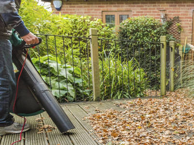 Eine Person nutzt einen elektrischen Laubsauger, um trockenes Laub von einer Holzterrasse vor einem Gartenzaun zu entfernen