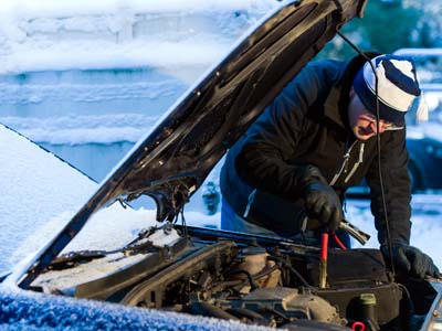 Foto von einem Mann, der in Winterkleidung und bei Schnee an einem Auto mit geöffneter Motorhaube steht und Starthilfekabel an der Batterie anschließt