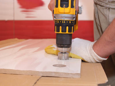 Ein Handwerker mit Arbeitshandschuhen bohrt mit einer gelb-schwarzen Bohrmaschine ein rundes Loch in eine einzelne Fliese. Die Fliese liegt auf Pappe, um Beschädigungen zu vermeiden