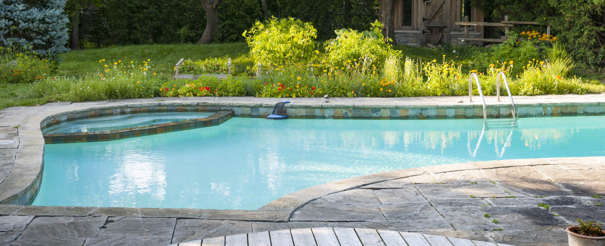 Ein in Naturstein eingefasster Gartenpool mit klarem, hellblauem Wasser. Im Hintergrund blühen bunte Stauden und Wiesenpflanzen, daneben stehen Bäume und eine kleine Holzlaube