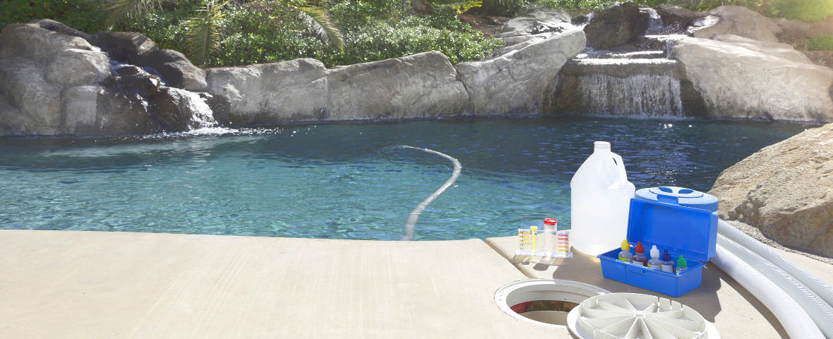 Ein sauberer Außenpool mit Wasserfällen im Hintergrund. Am Beckenrand stehen ein Wassertestset, eine Kanisterflasche und ein geöffneter blauer Kasten mit Chemikalien zur Poolpflege.