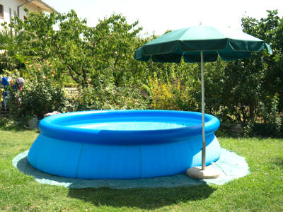 Ein runder, aufblasbarer Pool in kräftigem Blau steht auf einer Rasenfläche im Garten. Daneben befindet sich ein grüner Sonnenschirm mit hellem Standfuß, der Schatten spendet. Der Pool steht auf einer schützenden Unterlage, umgeben von Bäumen und Sträuchern