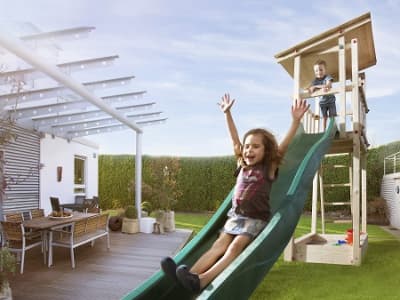 Zwei spielende Kinder auf einem Spielturm aus Holz mit einer grünen Rutsche, ein Mädchen rutscht und ein Junge steht oben auf dem Turm