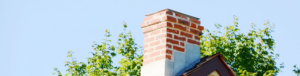 Ein gemauerter Schornstein mit Abdeckung ragt über das Hausdach und hebt sich deutlich vor blauem Himmel und Bäumen ab