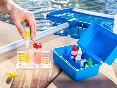 Ein Wassertestset zur Poolpflege wird am Beckenrand verwendet. Eine Hand tropft Reagenzflüssigkeit in eine Testvial. Daneben steht ein geöffneter blauer Kasten mit weiteren Prüfmitteln und Farbreferenzskala.