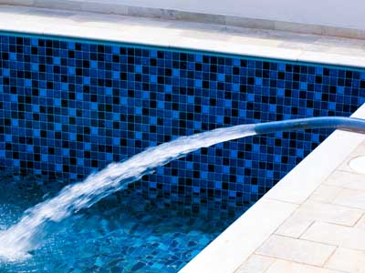 Ein mit blauen Mosaikfliesen ausgekleideter Pool wird über einen Schlauch mit Wasser befüllt. Der Schlauch liegt am Beckenrand auf hellem Boden.