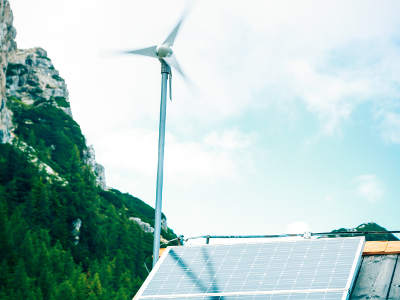 Ein Solarpanel und eine kleine Windturbine zur autarken Stromversorgung befinden sich auf dem Dach eines Gebäudes in den Bergen. Im Hintergrund sind Felsen und Wald zu sehen
